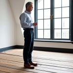 A professional interior designer, fully clothed in a modest business casual outfit, stands in the living room of a renovated old Dutch house, gently touching the surface of a wide, oiled oak plank floor. Sunlight streams through a large, authentic window, highlighting the rich texture and subtle imperfections of the vintage wooden planks. The room has high ceilings and tasteful, minimal decor that complements the historic architecture. The atmosphere is calm and inviting. safe for work, appropriate content, fully clothed, professional, perfect anatomy, correct proportions, natural pose, well-formed hands, proper finger count, natural body proportions, professional photography, high quality, Dutch heritage home.